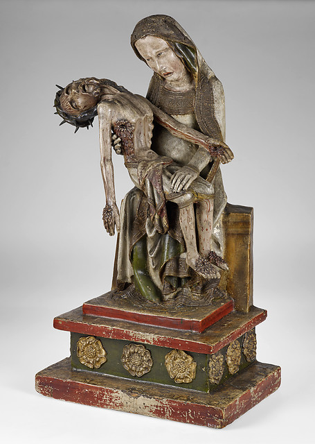 Painted wood sculpture of the Virgin Mary seated and holding the small, wounded body of the crucified Christ across her lap; both figures' faces show open grief.