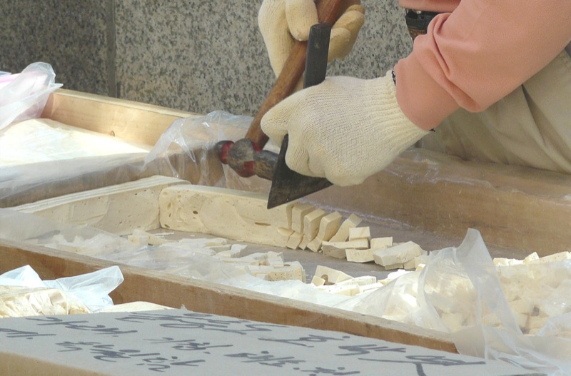 Gloved hands chiseling a long pale block of yeot into small squares on a wooden tray at a street stall, with finished pieces bagged in plastic alongside.