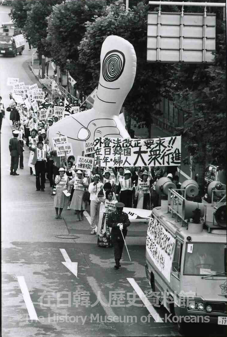 A 1984 street protest photographed from above: a long column of Zainichi Korean marchers in white caps carry placards beside a sound truck, led by a giant inflatable hand whose raised thumb bears a fingerprint whorl — a protest against the Alien Registration Act's fingerprinting requirement.
