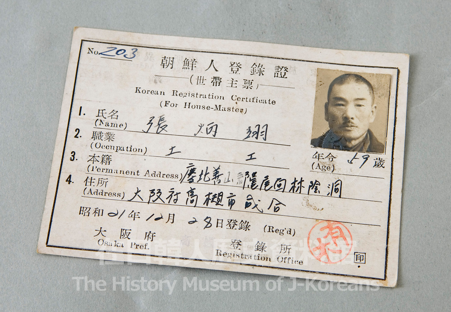 A 1946 Korean Registration Certificate (朝鮮人登録證) issued by Osaka Prefecture: a worn card printed in Japanese and English, hand-filled with the holder's name, occupation and address, bearing a sepia ID photograph of a man and a red official seal.