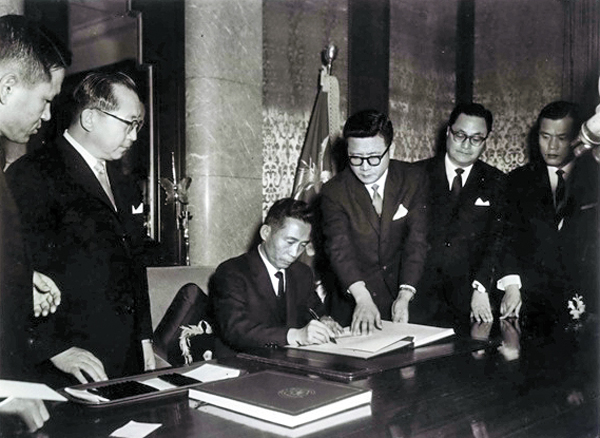 Black-and-white photograph of President Park Chung-hee seated at a desk signing the 1965 normalization treaty at the Blue House, flanked by officials in dark suits.