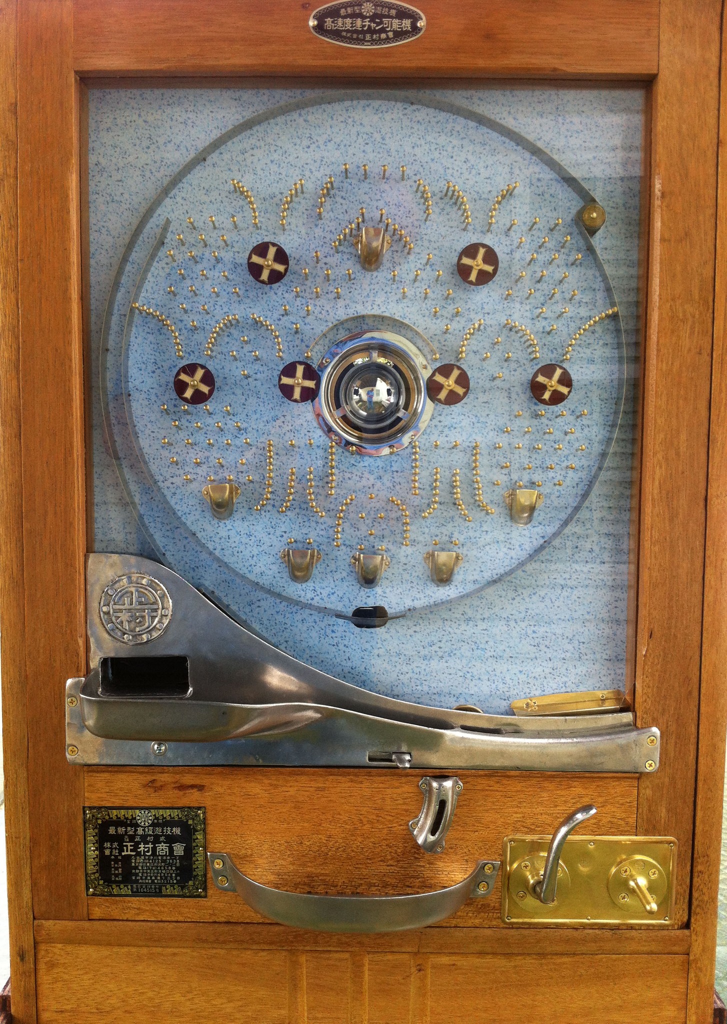 The face of a restored 1960s Shōraku pachinko machine: a pale blue speckled circular playfield studded with brass pins in fan patterns, maroon-and-gold spinner targets, brass tulip catchers, and a wood cabinet with a metal ball tray and brass launch handle.