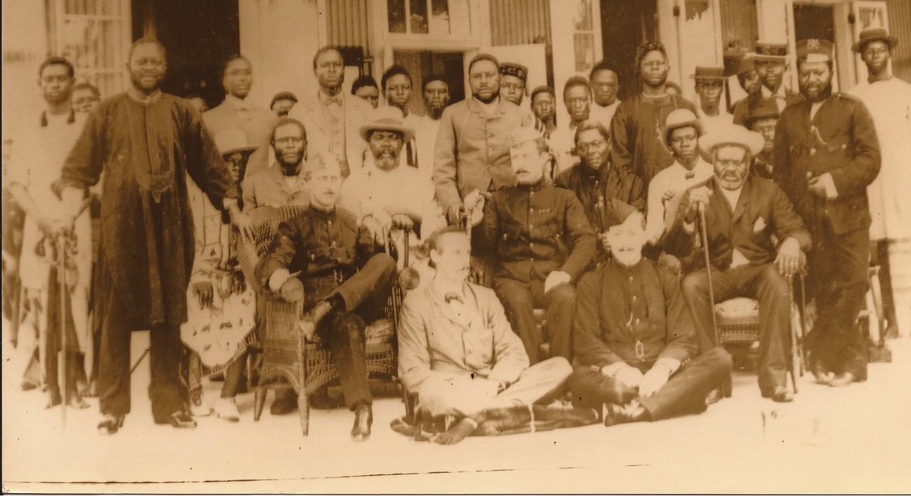 A sepia photograph from 1896: a large group of Bonny chiefs in formal dress posed on the veranda of a colonial building with the British Naval Commandant of the Bight of Biafra and Bight of Benin and other uniformed officials seated among them.