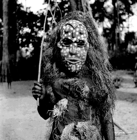 Five Ekpe masquerade figures standing in an outdoor clearing in southeastern Nigeria, wearing elaborate fiber costumes and painted masks. Photographed by G.I. Jones, c. 1930s.