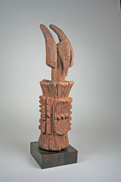 A carved wooden Igbo Ikenga shrine figure with two large upswept ram's horns rising from a stylized head, the body incised with rows of geometric notches, mounted on a dark display base.