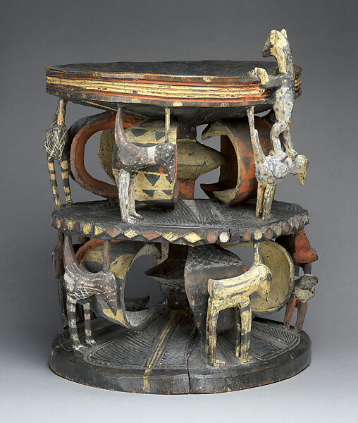 A two-tier carved wooden Igbo prestige stool with registers of concentric geometric patterns and standing animal figures around its supports, the surfaces painted in ochre, white, and dark pigments.